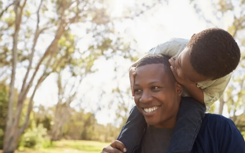 A father outdoors with his child on his shoulders, both laughing